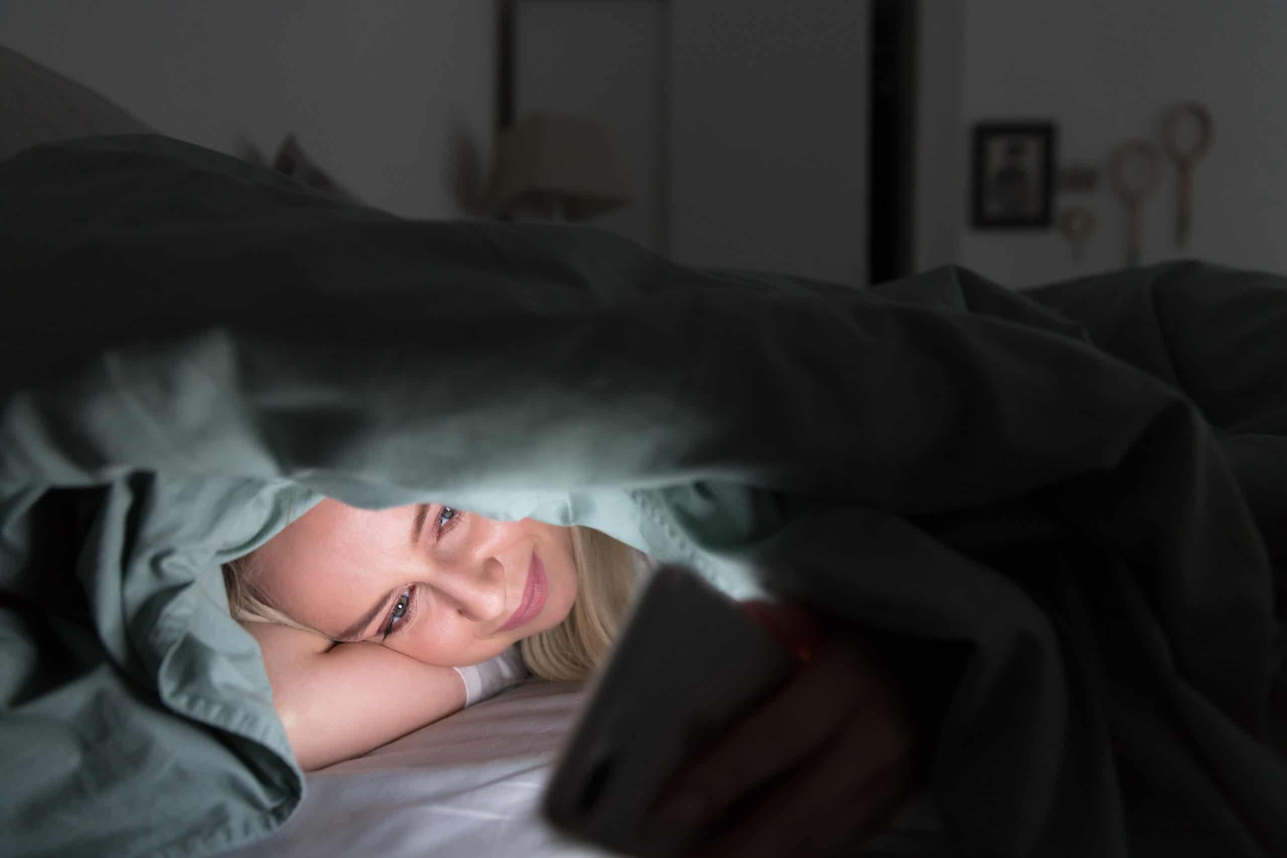 Junge Frau, die nachts mit Handy im Bett unter der Decke liegt, kann nicht schlafen
