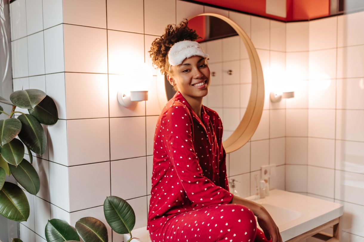 Frau in rotem Pyjama und Schlafmaske sitzt im Badezimmer und lächelt