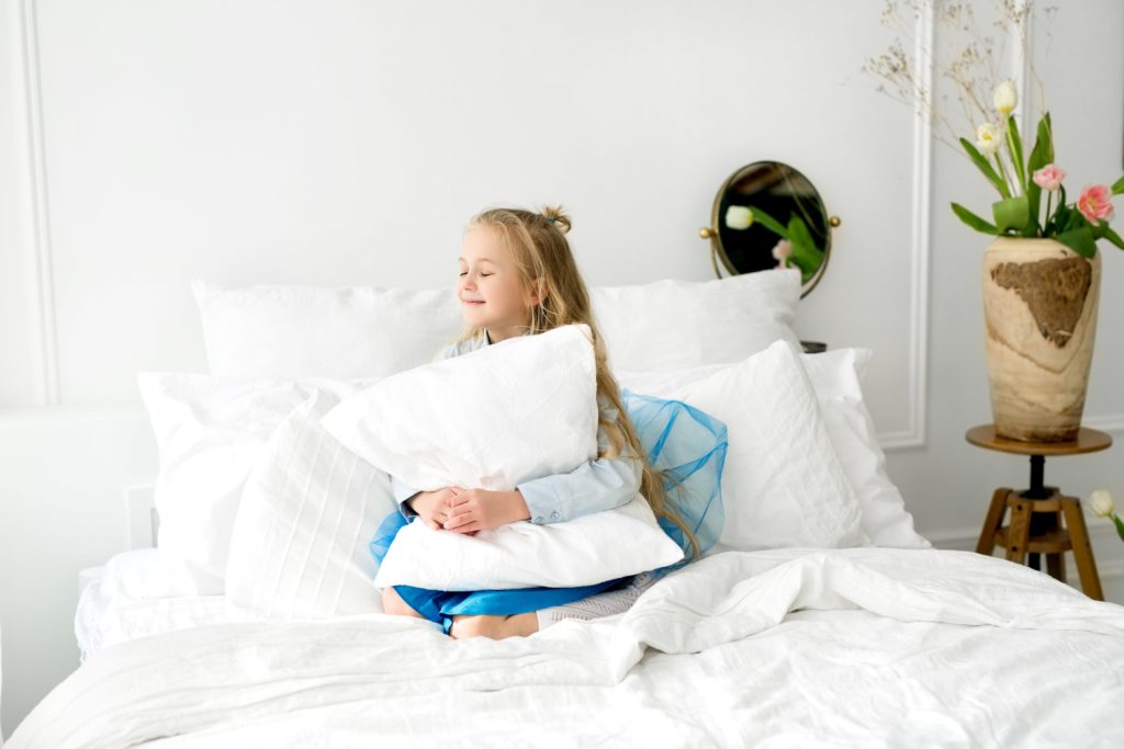 Mädchen sitzt in einem Bett mit schneeweißer Bettwäsche und umarmt ein Kissen
